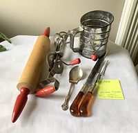 Photo displaying a wooden rolling pin with red handles, a metal flour sifter with red handle, a manual hand mixer with red handle, two-piece carving set with amber handles, and a silver-plated tomato spoon on a white tablecloth.