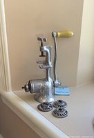 Full side view of vintage metal meat grinder showing clamp, crank with yellow handle, and one of the blades on the front.