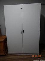 Closed front view of white wardrobe cabinet with two doors and black handles against wall.