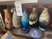 Photo of seven glass vases displayed on a wooden bench with varying colors including blue, yellow, pink, and multicolor swirl patterns. Two small decorative glass lids with iridescent finishes are in front.