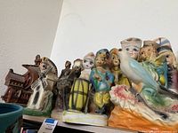 Wide view of multiple ceramic statues arranged on a shelf including a windmill, man figurines, busts, and horseman figure.