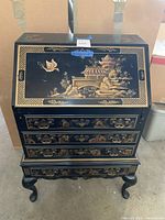 Full front view of the black wooden chest with gold etched Asian landscape design, drawers, and cabriole legs.
