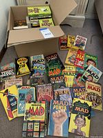 Photo shows an open box containing paperback books and a spread of various MAD magazine paperback editions and other books laid out on carpeted floor.
