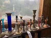 Photo showing 15 assorted candlesticks on a windowsill in natural light. Colors include blue, brass, silver, copper, pink, and greenish tones.