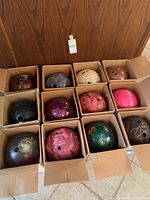 Twelve colorful bowling balls in individual cardboard boxes, showing a range of colors and patterns with three holes each.