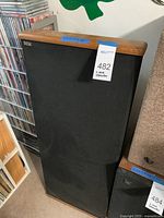 Front view of first DCM floorstanding speaker with oak top and black grille, lot tag visible