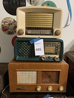 Front view of three stacked vintage radios: a cream GE radio on top, a green Silvertone radio in the middle, and a wooden cabinet radio with clock on bottom.