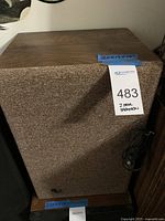 Front and top view of the Infinity speaker showing the wood veneer finish and fabric grille with label and blue tape indicating dimensions 20x13x10 inches.