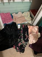 Multiple tops and skirts laid out in front of storage tub showing sequin mesh tops, crocheted top, checkered top, satin blouse, and velvet skirt