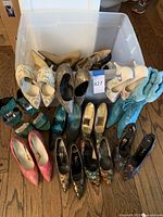 Wide view of lot shoes displayed on floor and bin, showing various colors and styles of high heels and boots including teal glitter, beige, snakeskin, floral embroidery, and fur boots.