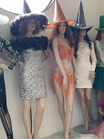 Four female mannequins dressed in costumes, featuring a silver dress with feather collar, beaded orange fringe outfit, white lace dress, and a seated mannequin holding a purple ball.