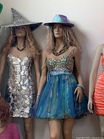 Two standing mannequins: one in silver sequin dress with witch hat, one in blue-green tulle dress with holographic hat