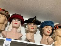 Top shelf overview of five mannequin heads displaying the fruit-adorned straw hat, red feather hat, net-veil black hat, blue velvet turban and striped-brim hat