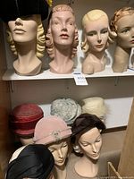 Four mannequin head busts on top shelf, four different hair colors and styles, one wearing black velvet pillbox hat