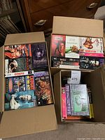 Three cardboard boxes filled with VHS tapes in used condition. Shows titles and cover art for various movies and instructional tapes.