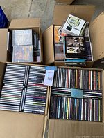 Four cardboard boxes filled with approximately a hundred music CDs by various artists, some stacked horizontally and others vertically with visible album titles.