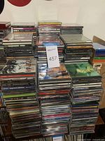 Multiple stacks of used music CDs in jewel cases with various artist and album titles visible, plus two small boxes of blank CD cases on the side.