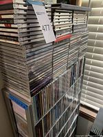 Photo showing tall stacks of music CD jewel cases on and around a transparent holder stand. Cases are arranged vertically and horizontally with visible album spines showing.