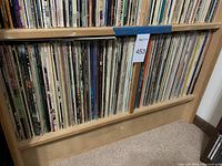 Wooden shelving unit filled with two rows of vinyl LP records showing spine views of numerous album jackets
