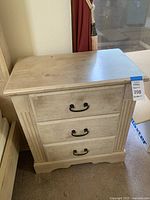 Full front view of the cream colored wooden side table showing three drawers with metal handles. Visible wear on bottom molding and drawer fronts.
