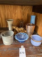 Photo showing a collection of 7 items on a wooden shelf: 2 ceramic vases, 3 ceramic bowls, 1 glass votive holder, and 1 gold decorative dog figurine.