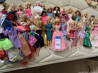 Photo showing a wide assortment of Barbie dolls in various dresses and casual wear, laid out on a couch with some loose clothes on the side.