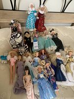 Photo showing a group of 17 vintage Barbie dolls, displayed on a bed, wearing a variety of costumes and gowns including blue fur-trimmed gown, red velvet dress, and jester outfit.
