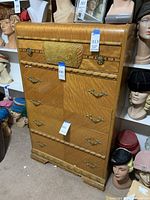 Full front view of the tall wooden dresser showing five drawers with gold tone pulls and carved details, surrounded by hats on mannequins