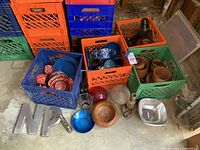Photo showing crates with red and blue enamel metal dishes, wooden decorative bowls and candle holders, large metal letters 'N' and 'P', and various small decor pieces.