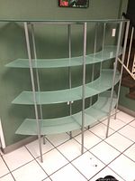 Photo of two modern curved shelving units with frosted glass shelves and silver metal frames, placed against a green wall on white tile floor. Each unit has three shelves with curved fronts.