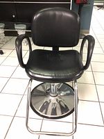 Frontal view of black vinyl salon chair showing padded seat and backrest with armrests, chrome round base with footrest.