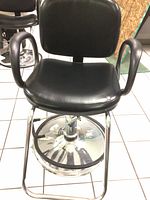 Front view of black vinyl salon chair showing padded seat, backrest, and armrests with round chrome base including footrest ring.