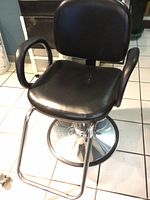 Full view of the black vinyl salon stylist chair showing padded seat, backrest, armrests, footrest, and round chrome base.