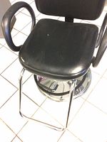 Front angled view of black vinyl seat and armrests attached to a chrome base on tiled floor. Minor wear visible on seat surface.