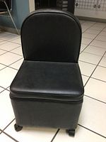 Front view of small black vinyl chair with padded seat and backrest, showing smooth upholstery and four caster wheels underneath.