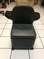 Front view of the black vinyl small chair with a moderate backrest and wheels on a white tile floor.