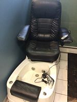 Full front view of black faux leather pedicure chair mounted on white acrylic foot spa basin with remote on armrest