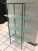 Front and side angle of curved frosted glass four-shelf unit with metal frame on tiled floor near a wall outlet.