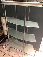 Full view of the glass shelving unit showing the four curved frosted glass shelves with a silver frame from the side and front.