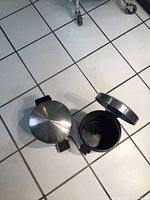 Two small aluminum pedal trash cans, one closed and one with lid open, on white tiled floor