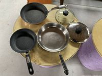 Three frying pans and two glass lids on a round surface: two pans with Teflon coating and one stainless steel pan, two glass lids with black knobs.