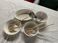 Set of four white bowls, three rounded ceramic, one oval serving dish, accompanied by four wooden spoons and a metal whisk with black handle, arranged on white cloth surface.