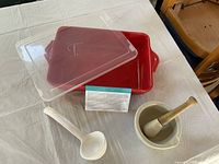 Red Tara casserole dish with clear lid and warranty card beside white ceramic mortar and pestle and soup spoon on a white cloth.