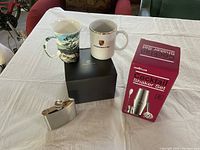 Photo showing two coffee cups, one Porsche branded in original box, a stainless steel flask, and an unopened cocktail shaker set box on a table.