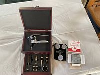 Open wooden box displaying silver executive style wine opener and additional wine accessories including foil cutter, pourers, and stoppers. Two sets of whisky stones visible: black pouch labeled The Glenlivet with six stones, and opened box labeled Cheers Canada with six stones.