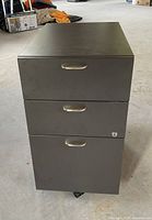 Front view showing three drawers with metal handles, lock on the middle drawer, and caster wheels.