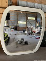 Photo of large white framed mirror leaning against a wooden structure inside a warehouse. Shows the mirror surface reflecting items in the background.