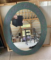 Oval mirror with decorative turquoise and purple mosaic-style frame, placed leaning against a wall in a workshop or storage space.