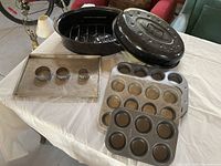 Photo showing two metal muffin tins (12-cup and 6-cup), a rectangular metal cookie sheet, and a large black enamel roasting pan with lid and internal rack on a table.