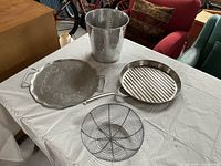 Four silver-colored items displayed on a table including a hammered metal waste basket, hammered tray, Calphalon ridged fry pan, and wire basket.